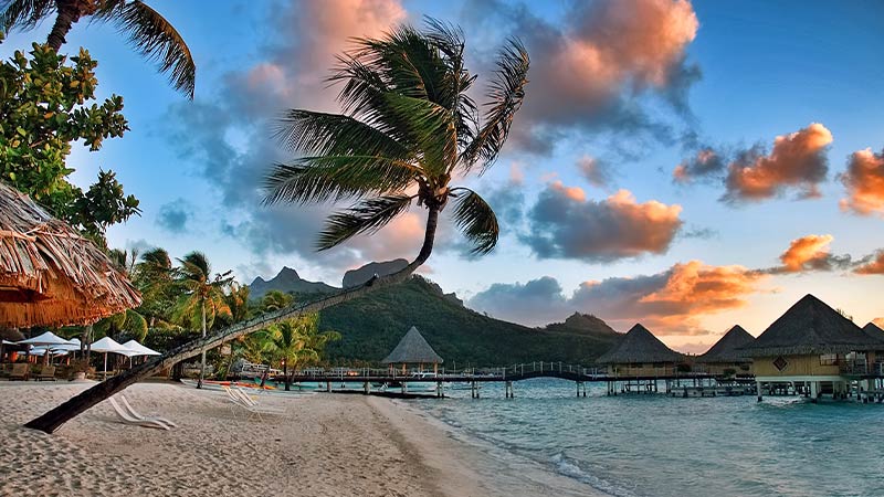 A beach in Bora Bora at golden hour with over the water bungalows in the background.