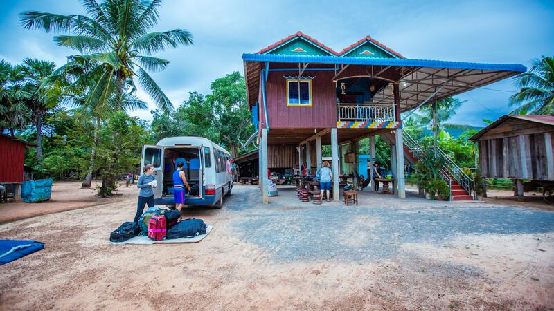 Two travellers unpack bags from a van that is parked in front of a colourful house on stilts. The house is painted red, green and blue and surrounded by palm trees.