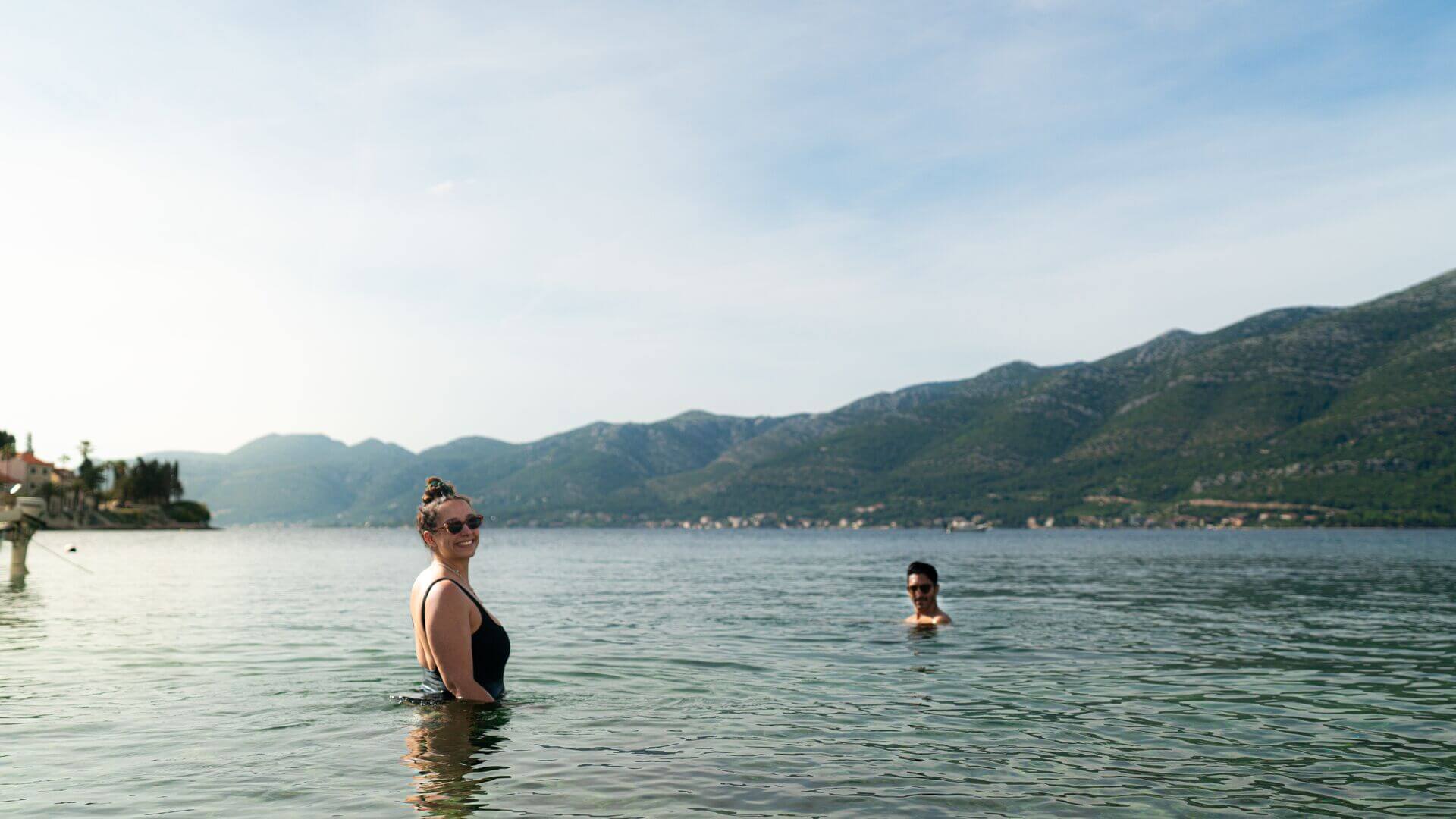 Two swimmers stand in the blue water with rolling green hills behind them at Korčula Island, Croatia