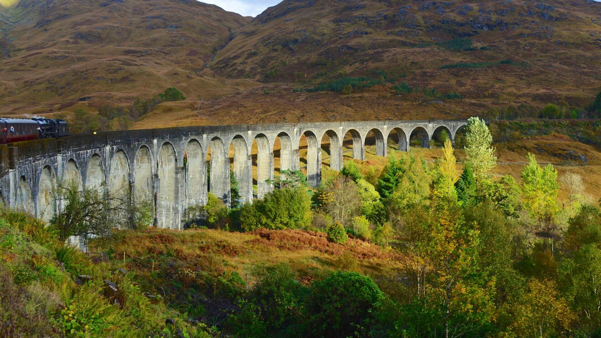 A steam train going across the Glenfinnan Viaduct with bright orange, green and red foliage, Scotland