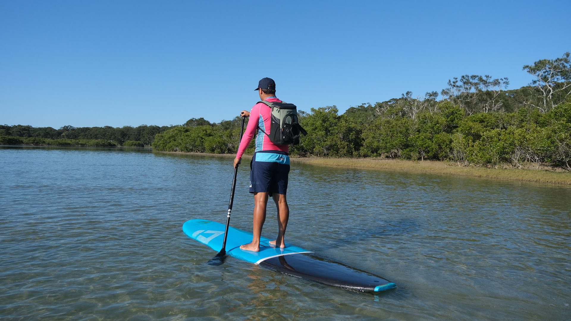 A paddle boarder wearing a pink sun shirt and blue shorts paddles on a calm creek