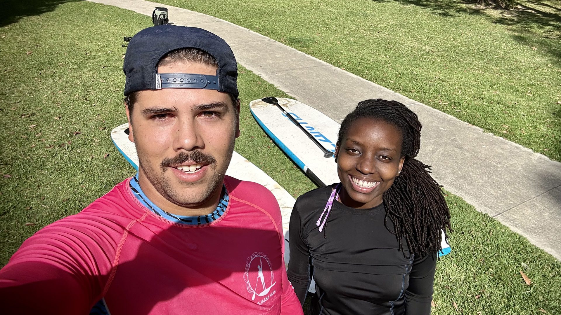 A stand up paddle board guide wearing a pink sun shirt takes a selfie with a traveller and their paddleboards.