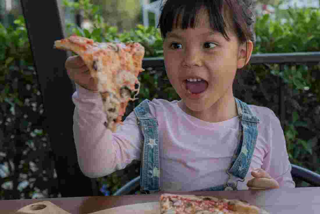 A young girl holding up a piece of pizza in Italy with her mouth open 